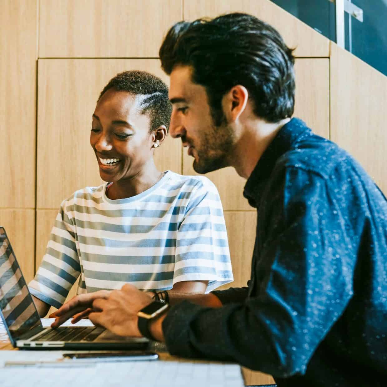 Two colleagues smiling while working together at a laptop in a modern office setting, with a coffee cup and notepad on the table.