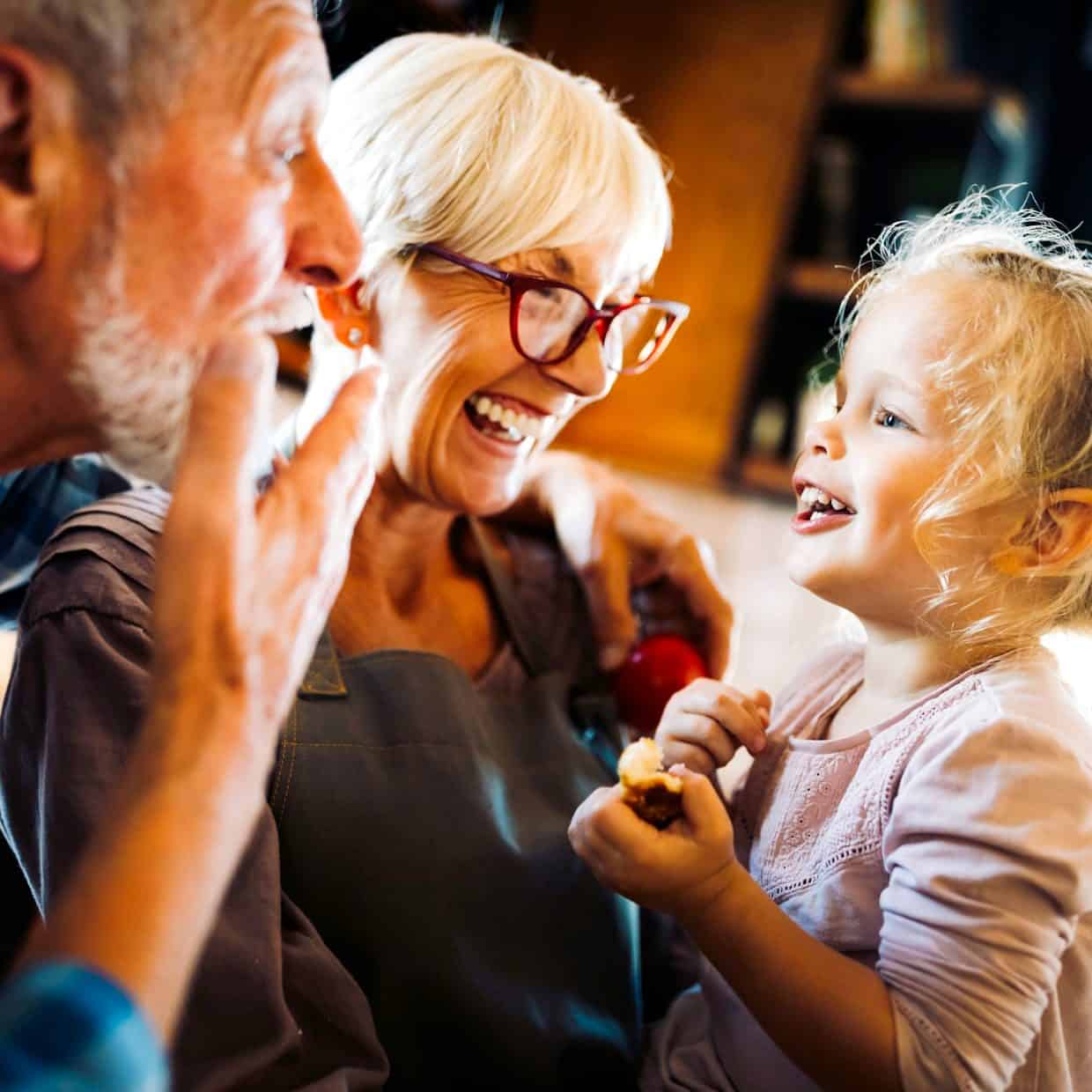 Happy grandparents laughing with their young granddaughter in a bright kitchen.