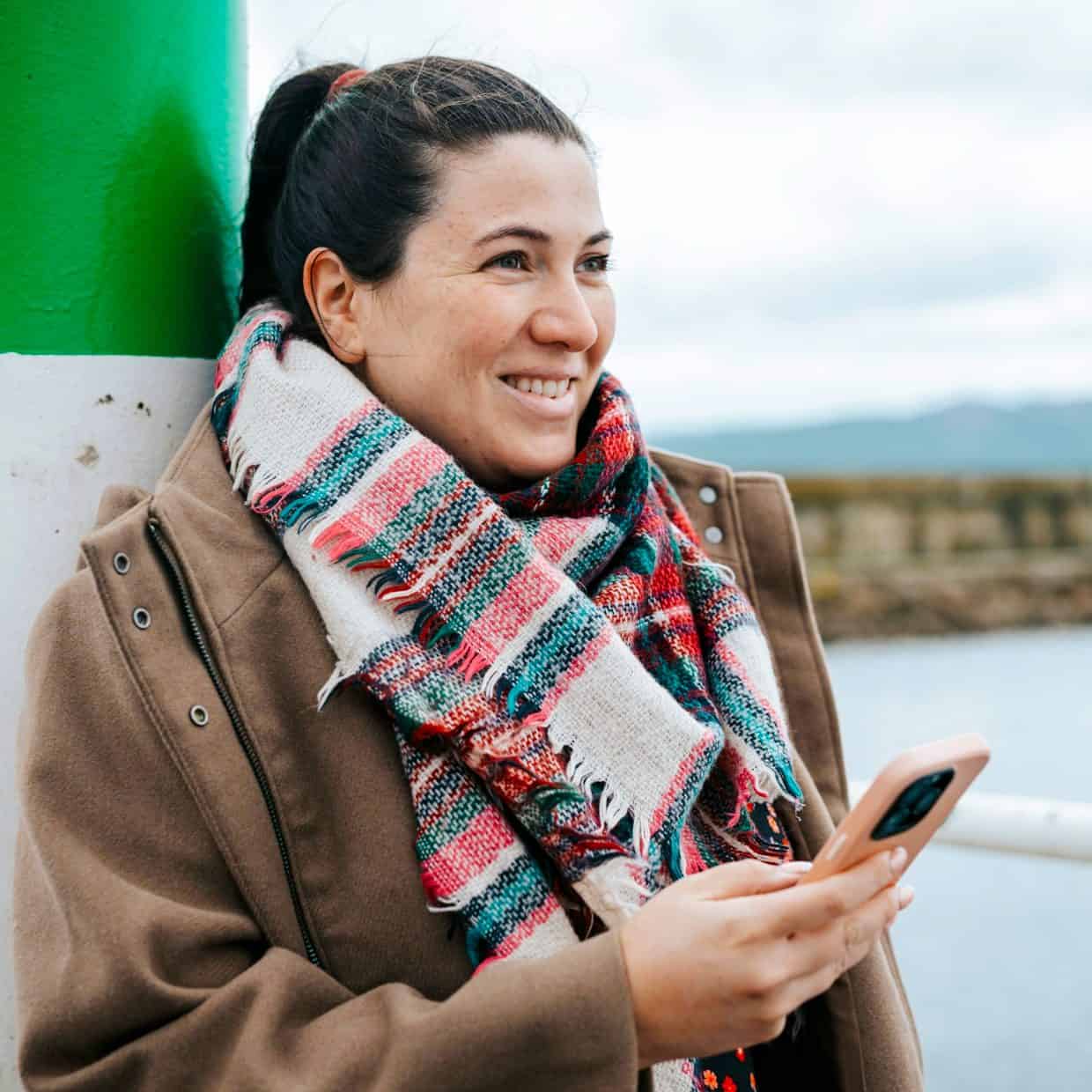Smiling woman in scarf using phone near water on a cold day.