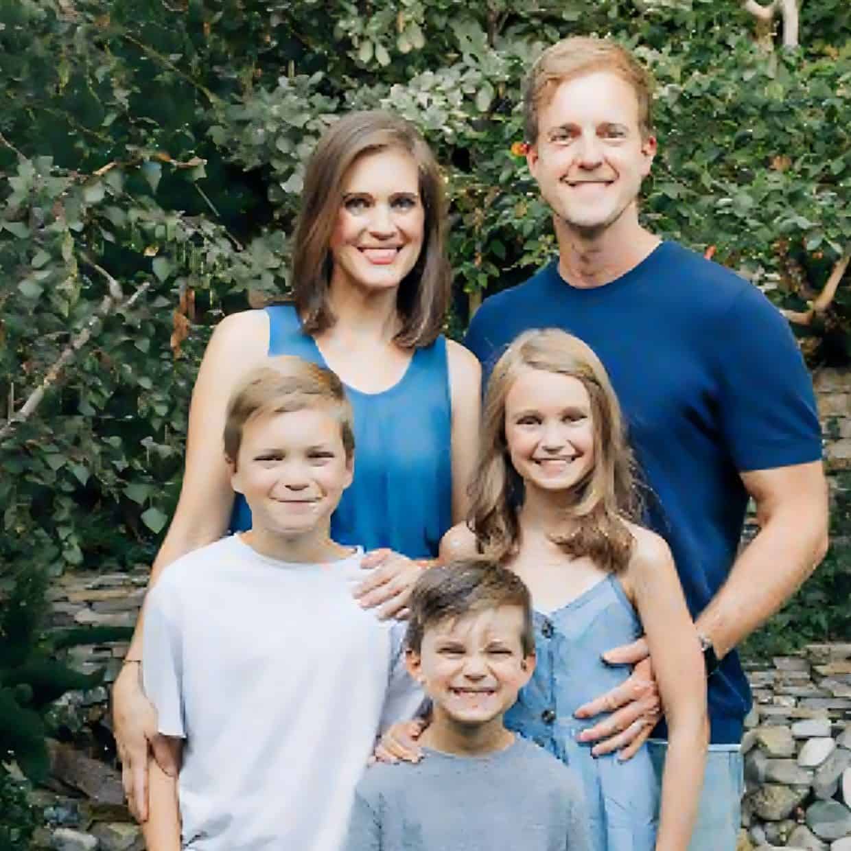 Smiling family of five posing outdoors in front of green foliage, with parents standing behind and three children in front.