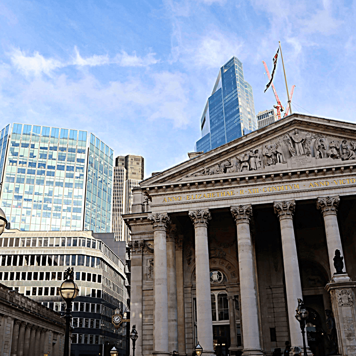 Historic Bank of England building with classical columns in the foreground, surrounded by modern glass skyscrapers in the City of London under a blue sky.