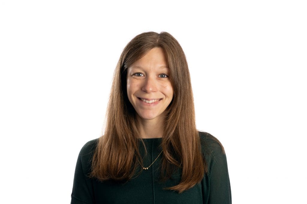 Smiling woman with long brown hair wearing a dark green top, photographed against a plain white background.