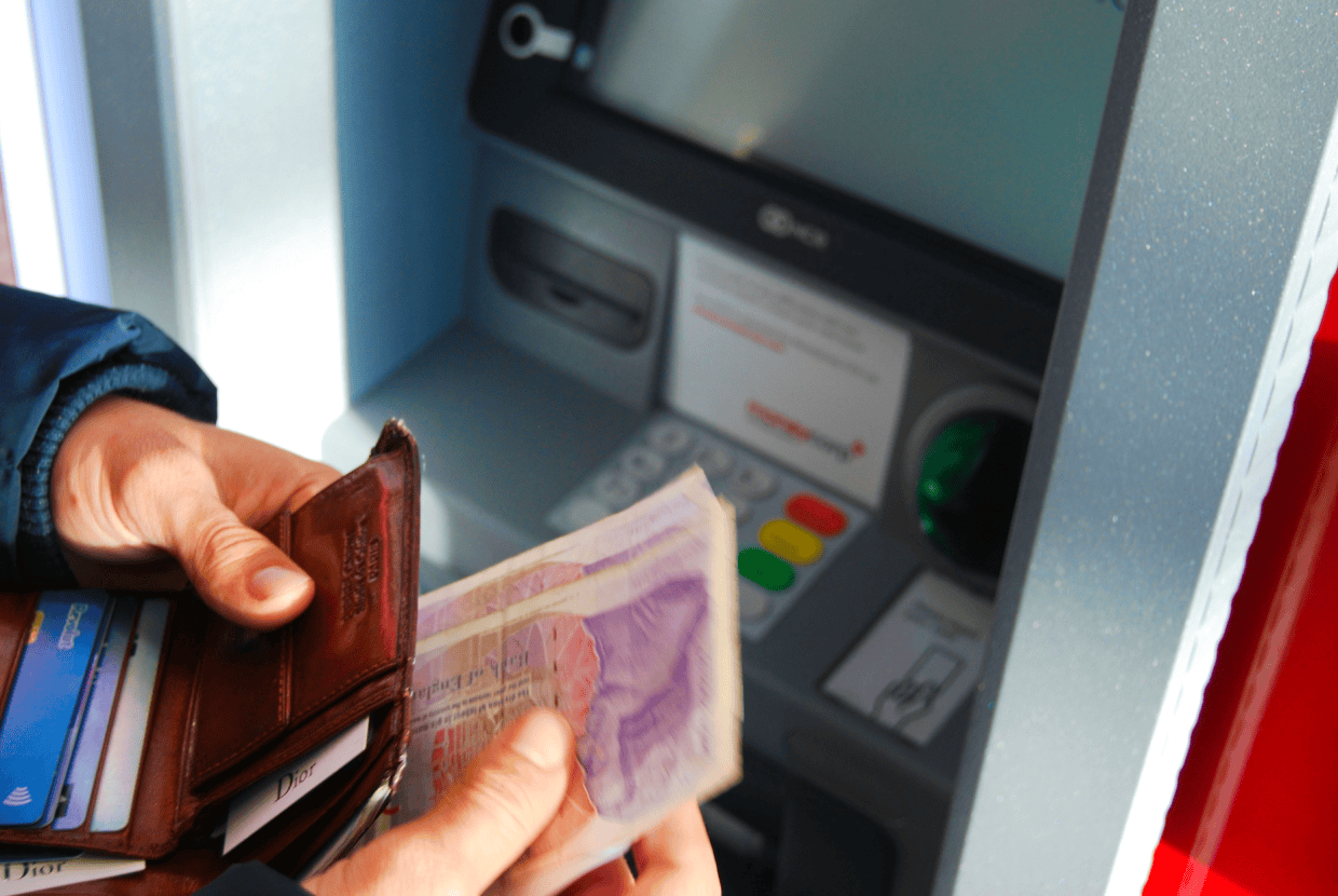 Person holding a wallet and banknotes while standing at an ATM machine, with card slots and keypad visible in the background.