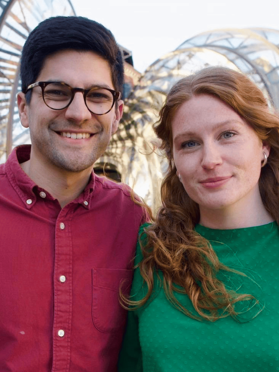 John and Imogen standing side by side outdoors, smiling at the camera with a modern arched structure and buildings in the background.