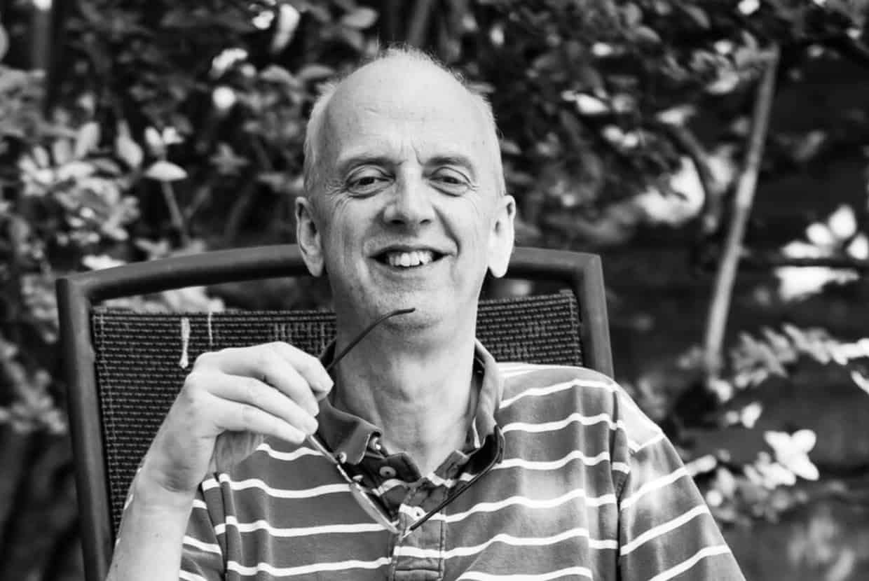 Jeremy Marshall smiling while holding glasses, seated outdoors with trees in the background.