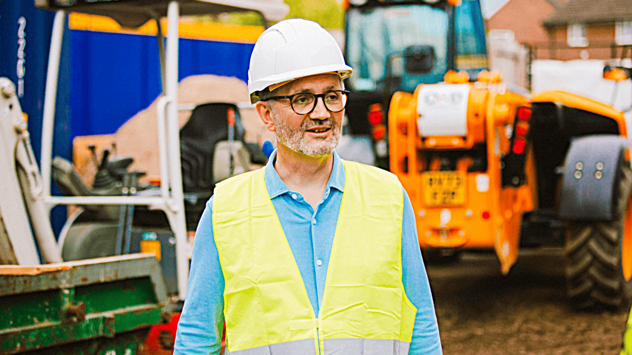 Jon Broome wearing a hard hat and high visibility vest standing at a construction site with machinery in the background.
