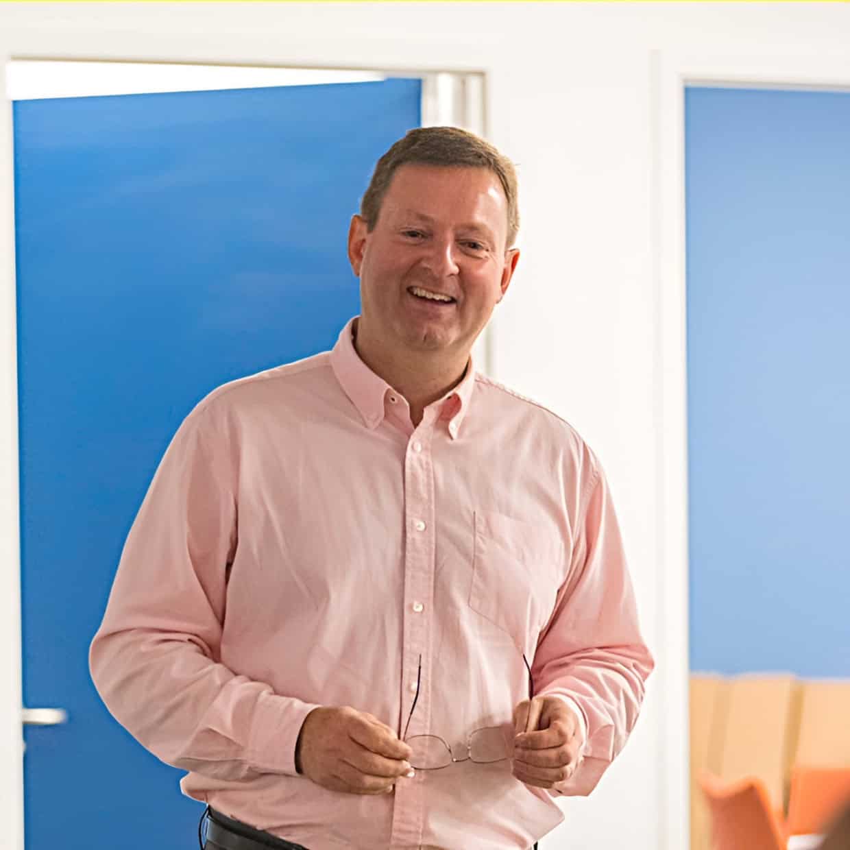 Paul Houghton smiling while speaking indoors, holding glasses in his hands with a bright background behind him.