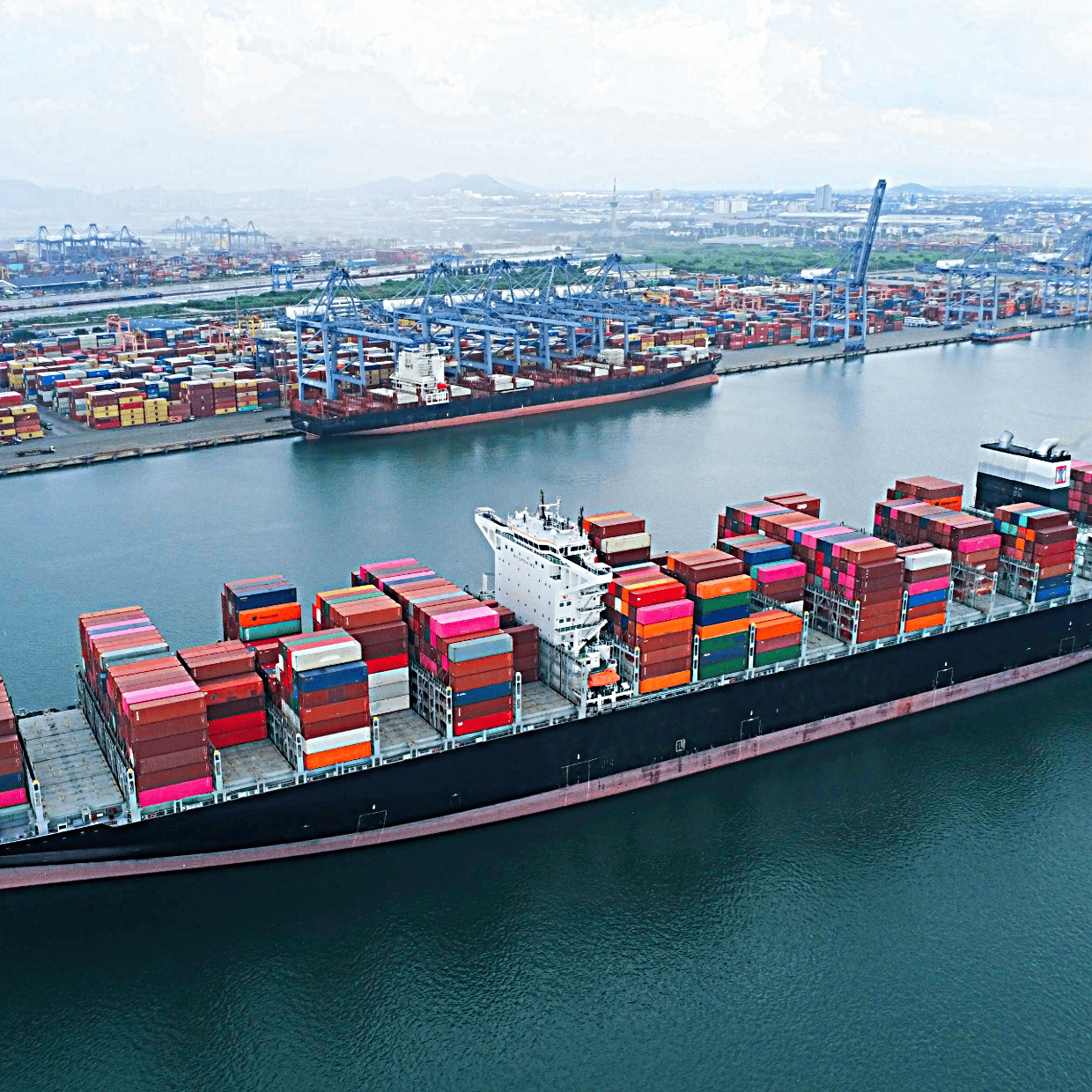 Large container ship carrying multicoloured cargo containers sailing past a busy commercial port with cranes and stacked containers.