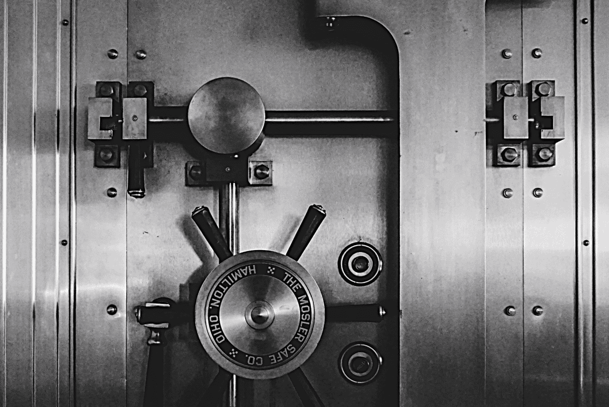 Close up of a heavy steel bank vault door with locking mechanism and wheel handle in black and white.