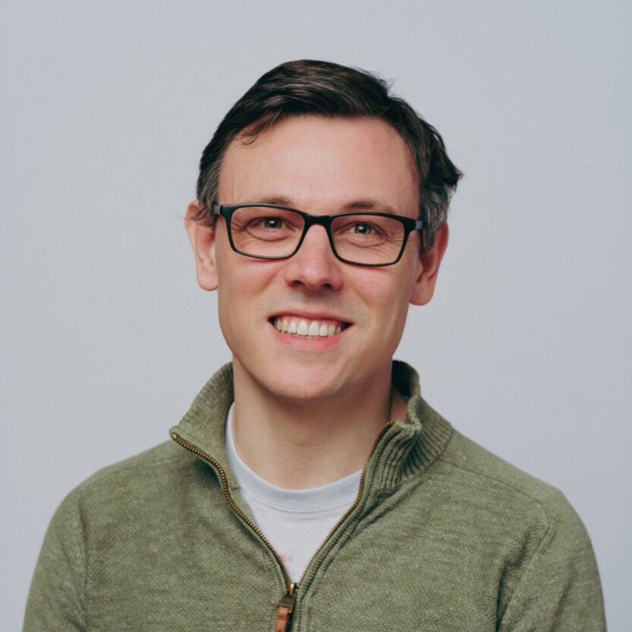 Smiling man wearing glasses and a green zip-neck jumper, standing against a plain light background.