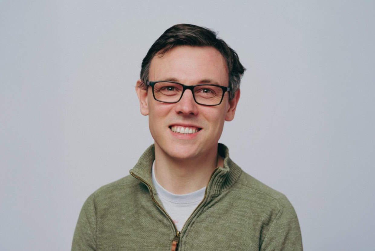 Smiling man wearing glasses and a green zip-neck jumper, standing against a plain light background.