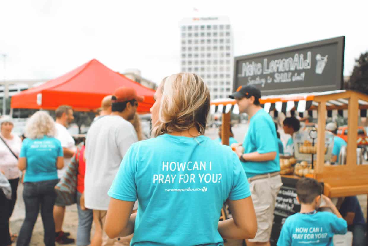 Person wearing a blue t-shirt reading “How can I pray for you?” at a busy outdoor market stall, with people gathered around in the background.