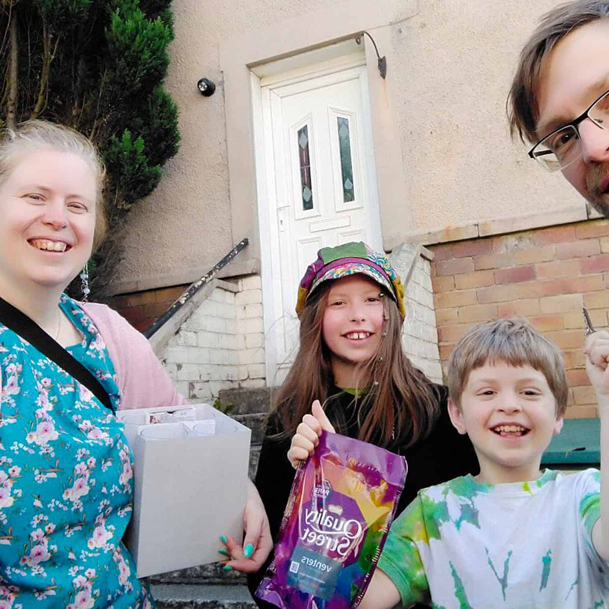 Smiling family of four standing outside their home, with the mother holding a box and the children holding sweets, celebrating with excitement.