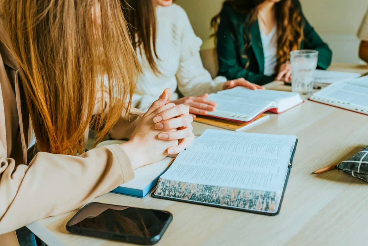 Group of people gathered around a table with open Bibles, studying scripture together, one person clasping their hands in prayer.