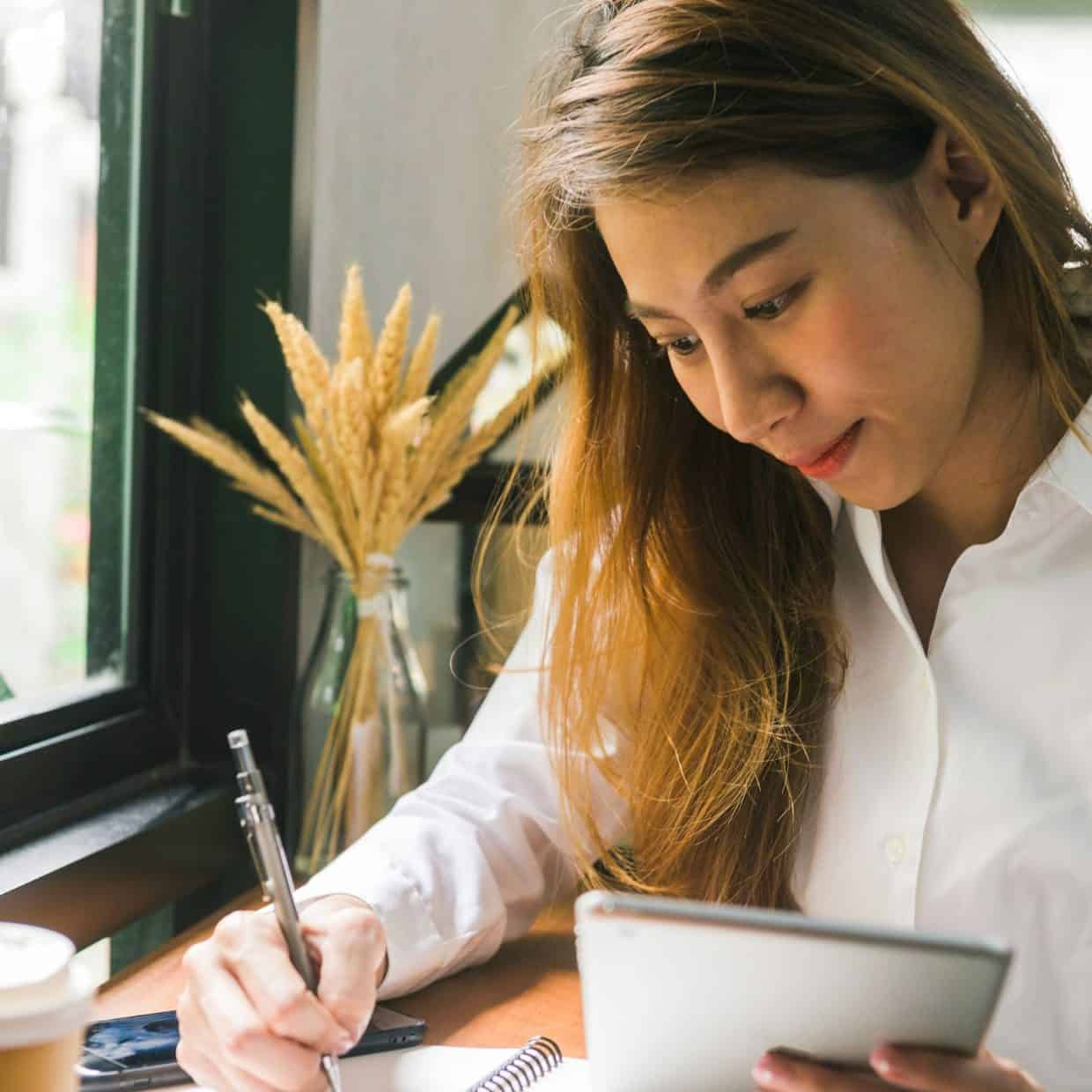 Young business woman writing in a notebook while holding a tablet by a window with coffee nearby.