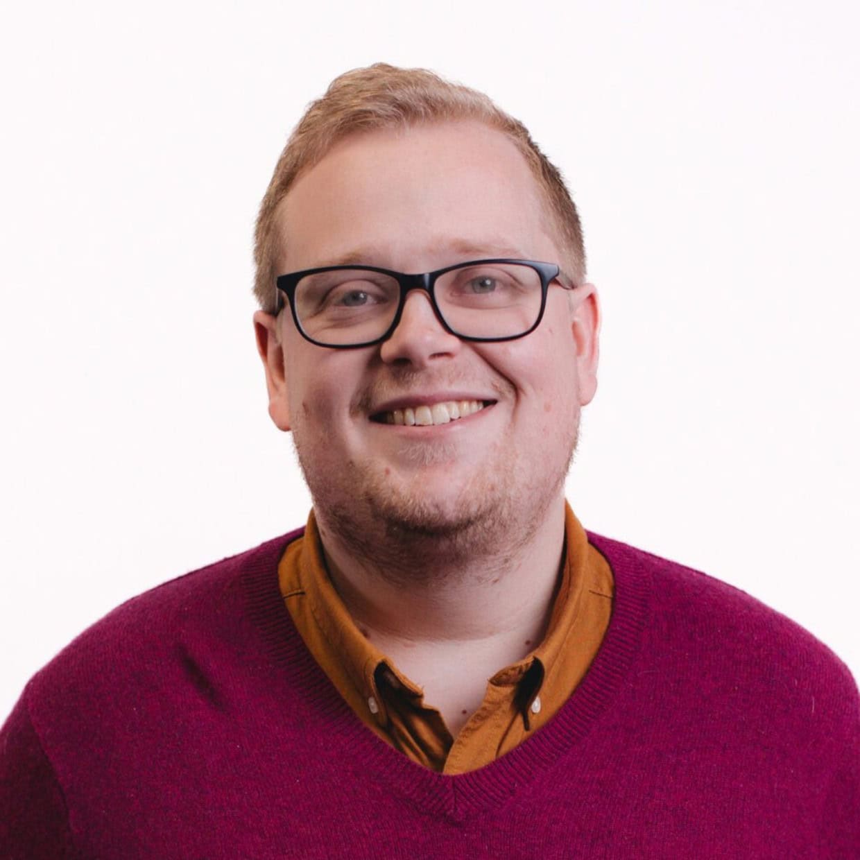 Smiling man wearing glasses, a mustard shirt, and a burgundy jumper, standing against a plain white background.