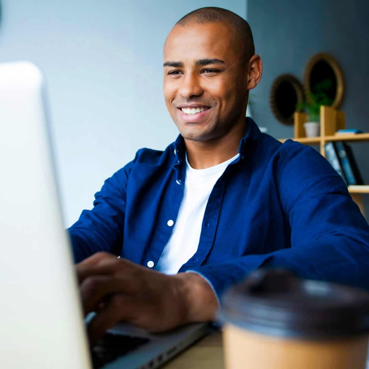 Man smiling while working on a laptop at a desk with a coffee cup nearby.