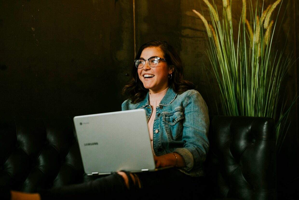 Smiling woman in glasses and a denim jacket using a laptop while sitting on a black leather sofa next to a tall plant.