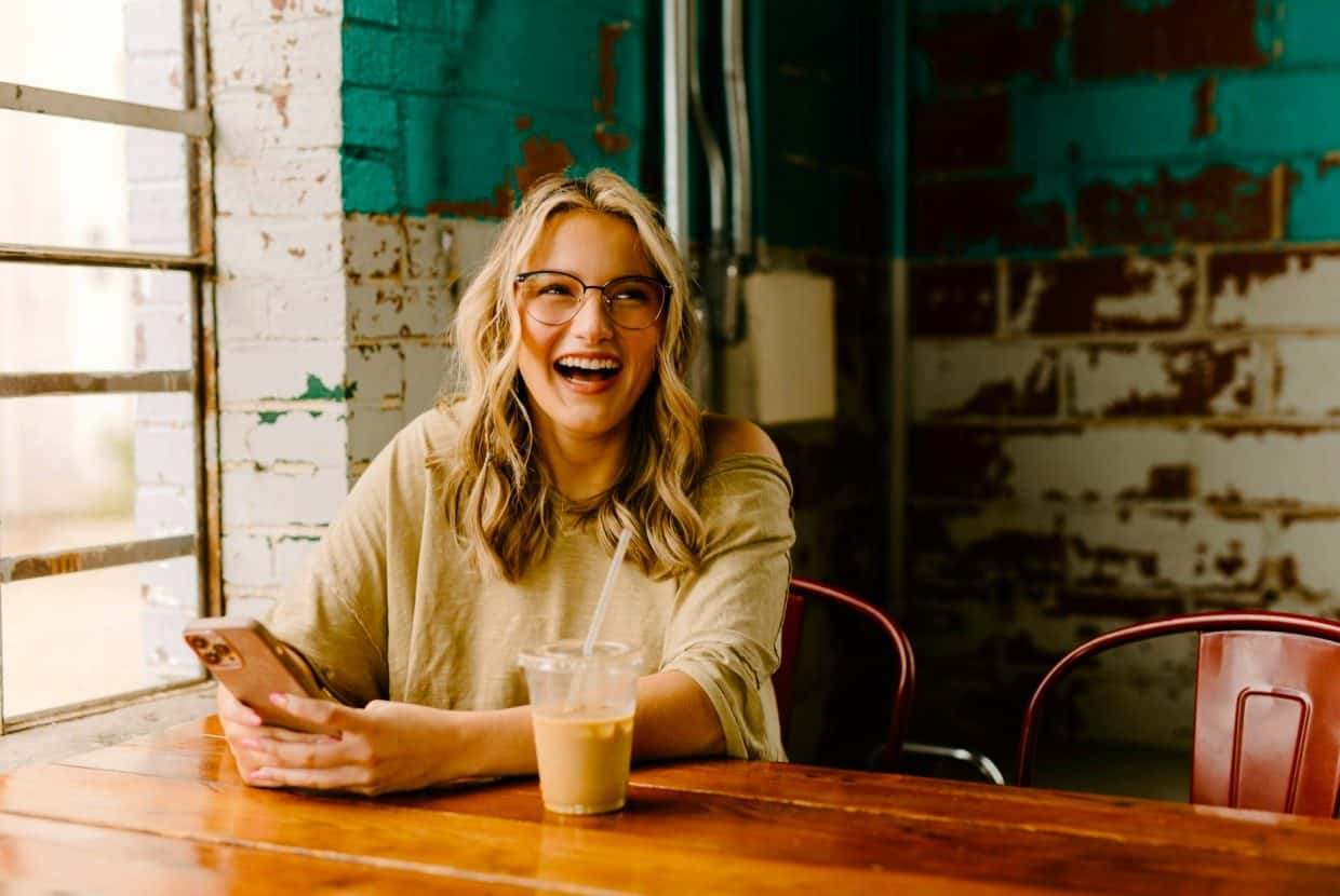 Smiling woman holding a phone and iced coffee while sitting in a rustic café.