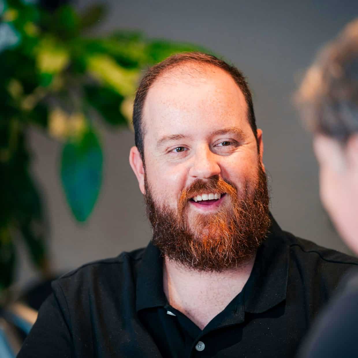 Bearded man in a black polo shirt smiling while talking to another person indoors.