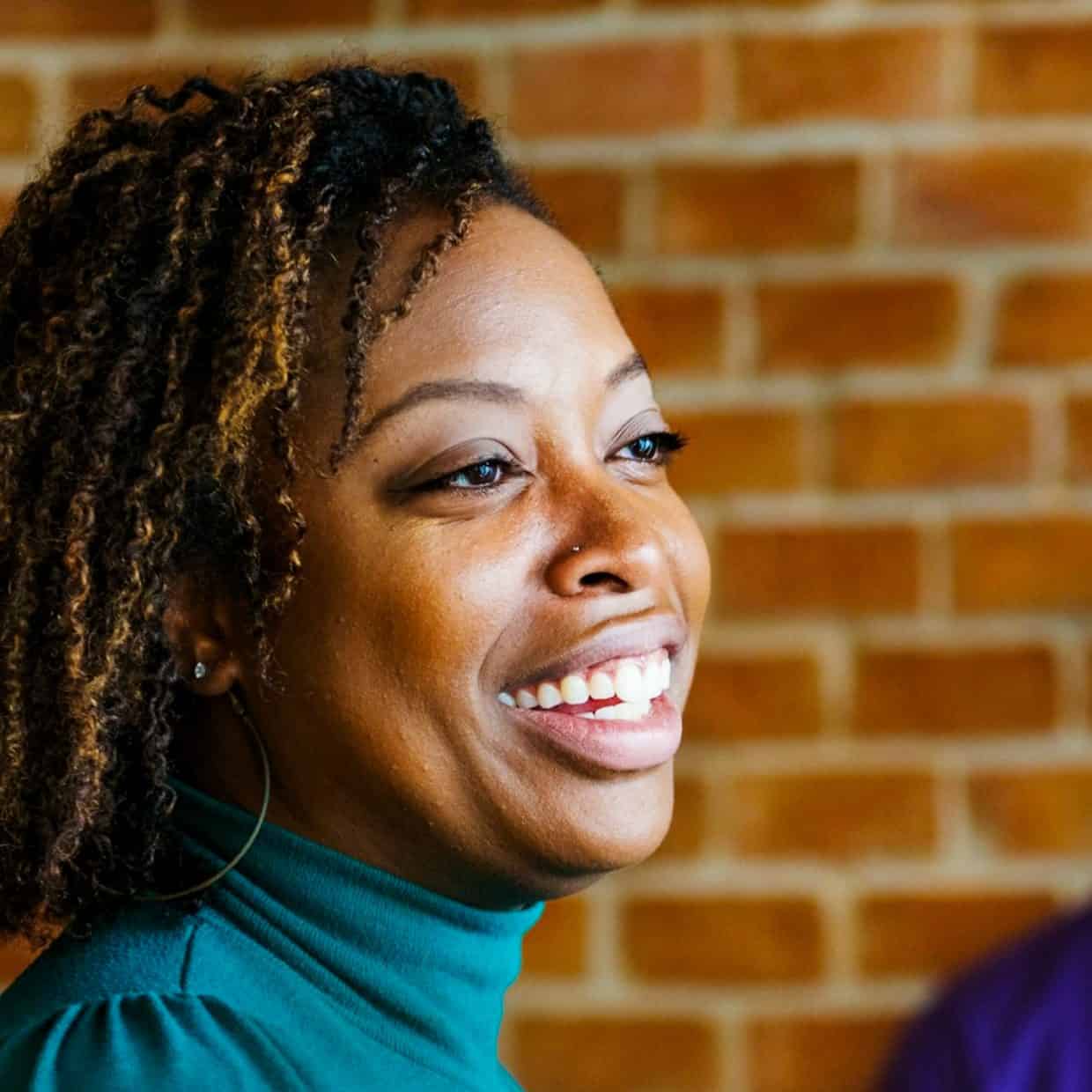 Smiling woman in a teal top with a brick wall background.