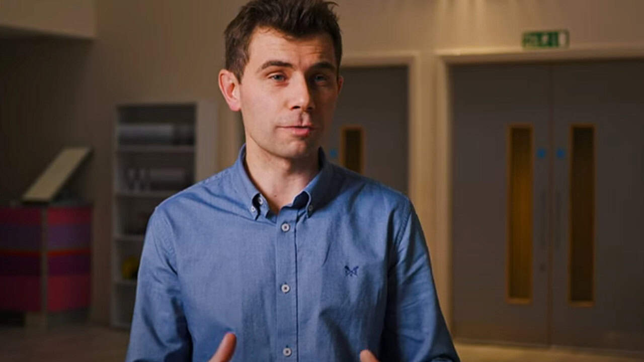 Man wearing a blue shirt speaking indoors, gesturing with his hands in front of closed double doors and soft lighting.
