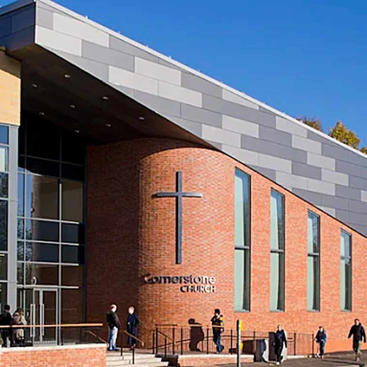 Modern red-brick building with large windows and a cross on the wall, showing the exterior of Cornerstone Church on a sunny day.
