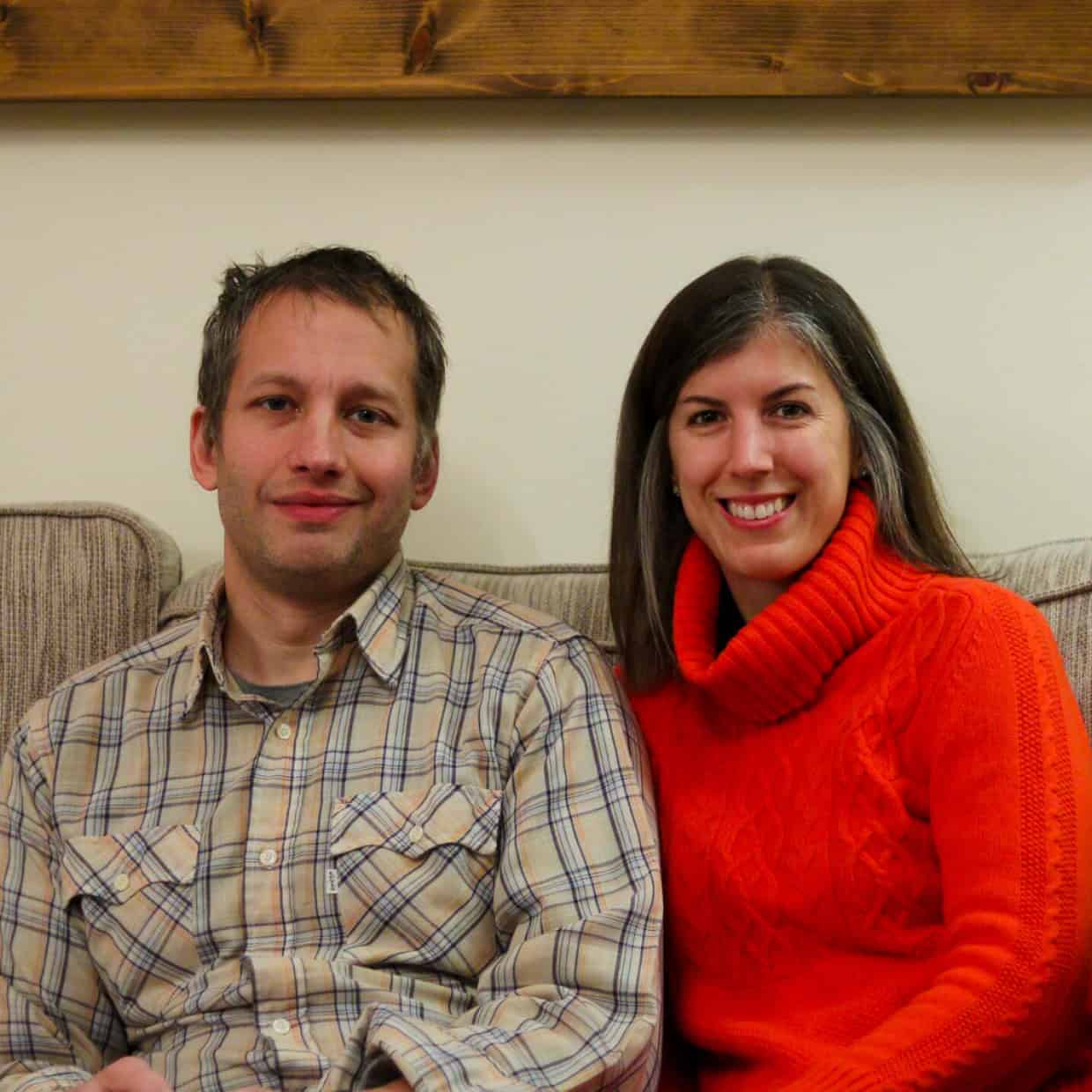 Man in a checked shirt and woman in a bright orange jumper sitting together on a beige sofa, smiling at the camera.