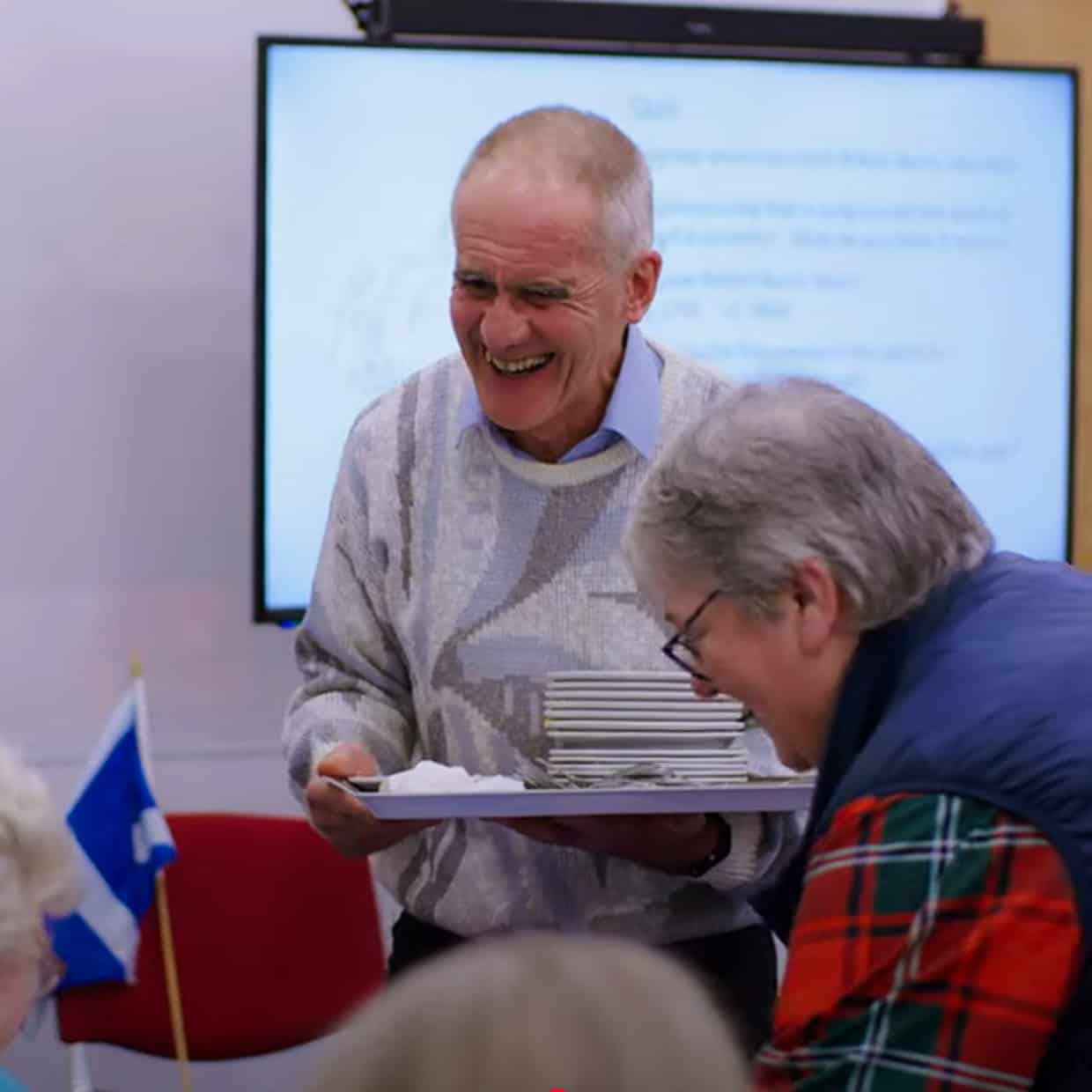 Smiling man holding a tray of plates while serving at a community event, with others seated and chatting in the background.