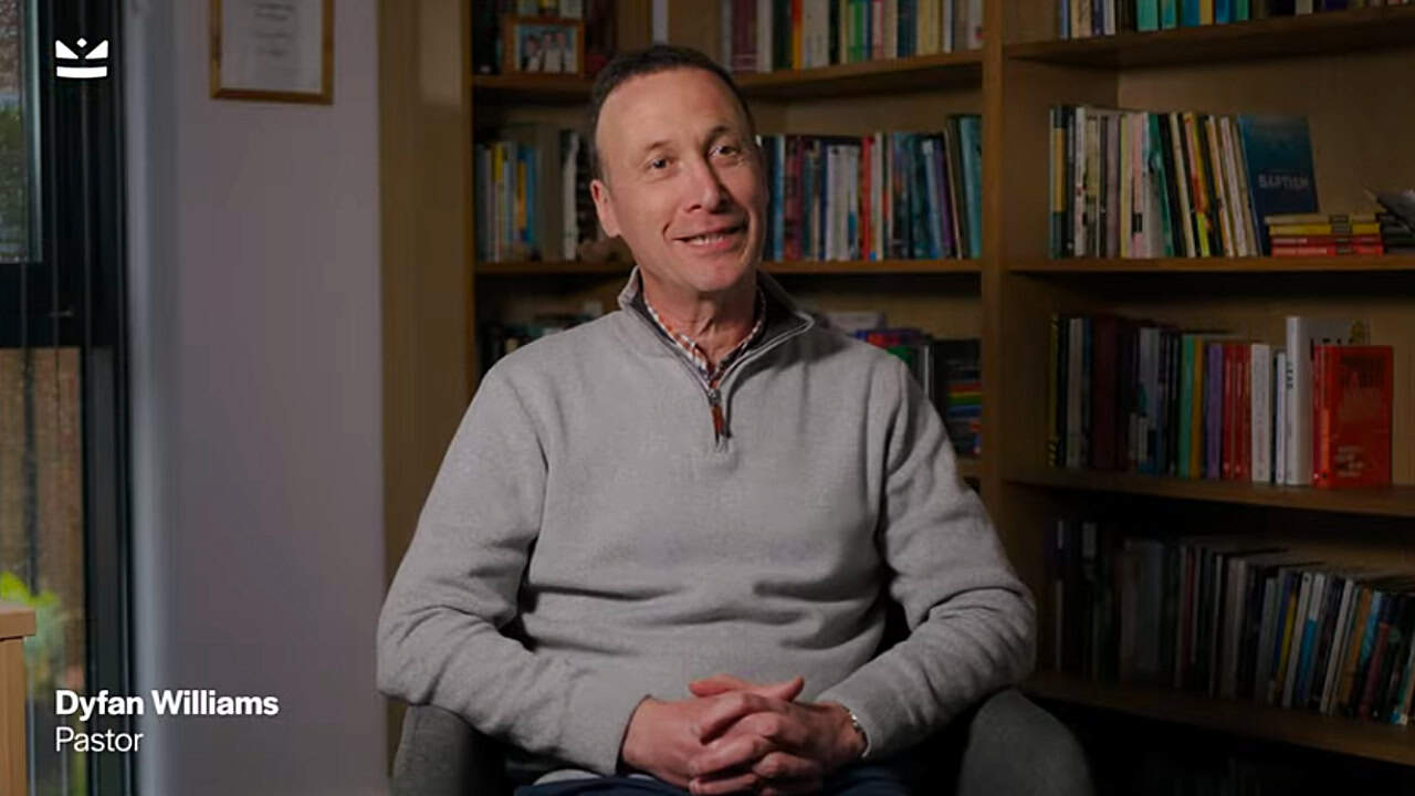 Man sitting and smiling in a chair in front of bookshelves filled with books, with text overlay in the corner reading 'Dyfan Williams, Pastor'.