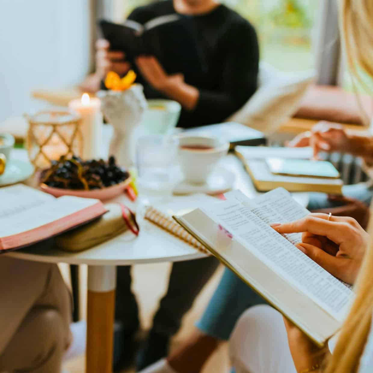 A group of people sitting around a table reading Bibles, with coffee, fruit, and candles on the table, suggesting a relaxed Bible study setting.