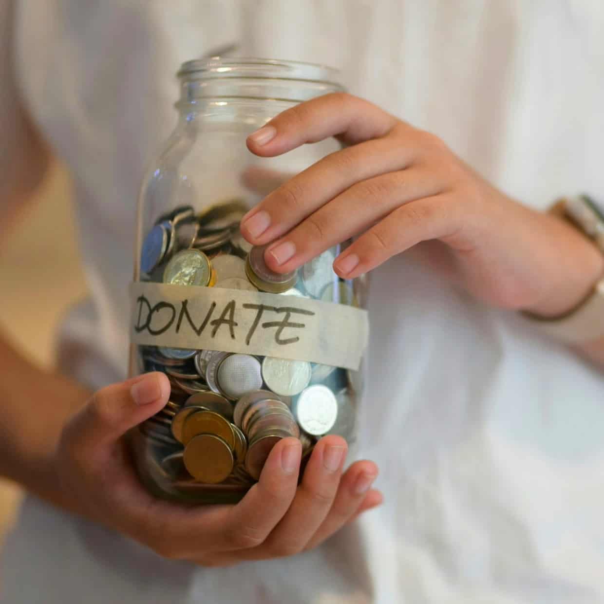 Person holding a glass jar filled with coins labeled 'Donate'.