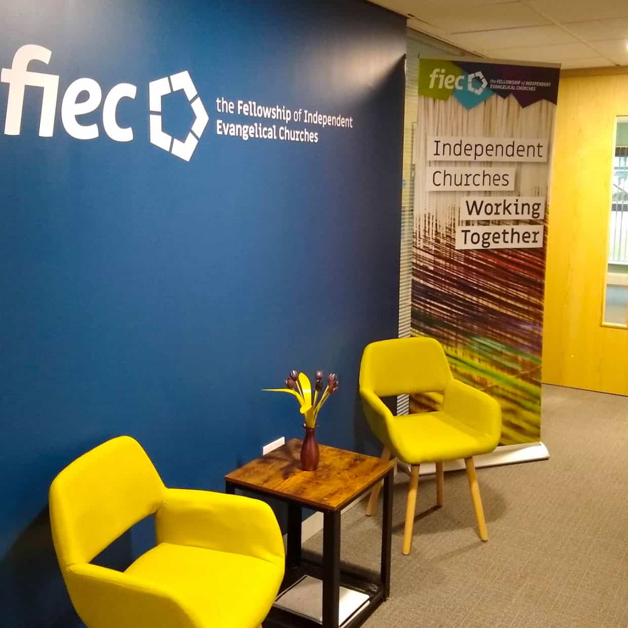 Modern office reception area with two yellow chairs, a small table, and a blue wall displaying the Fellowship of Independent Evangelical Churches logo.