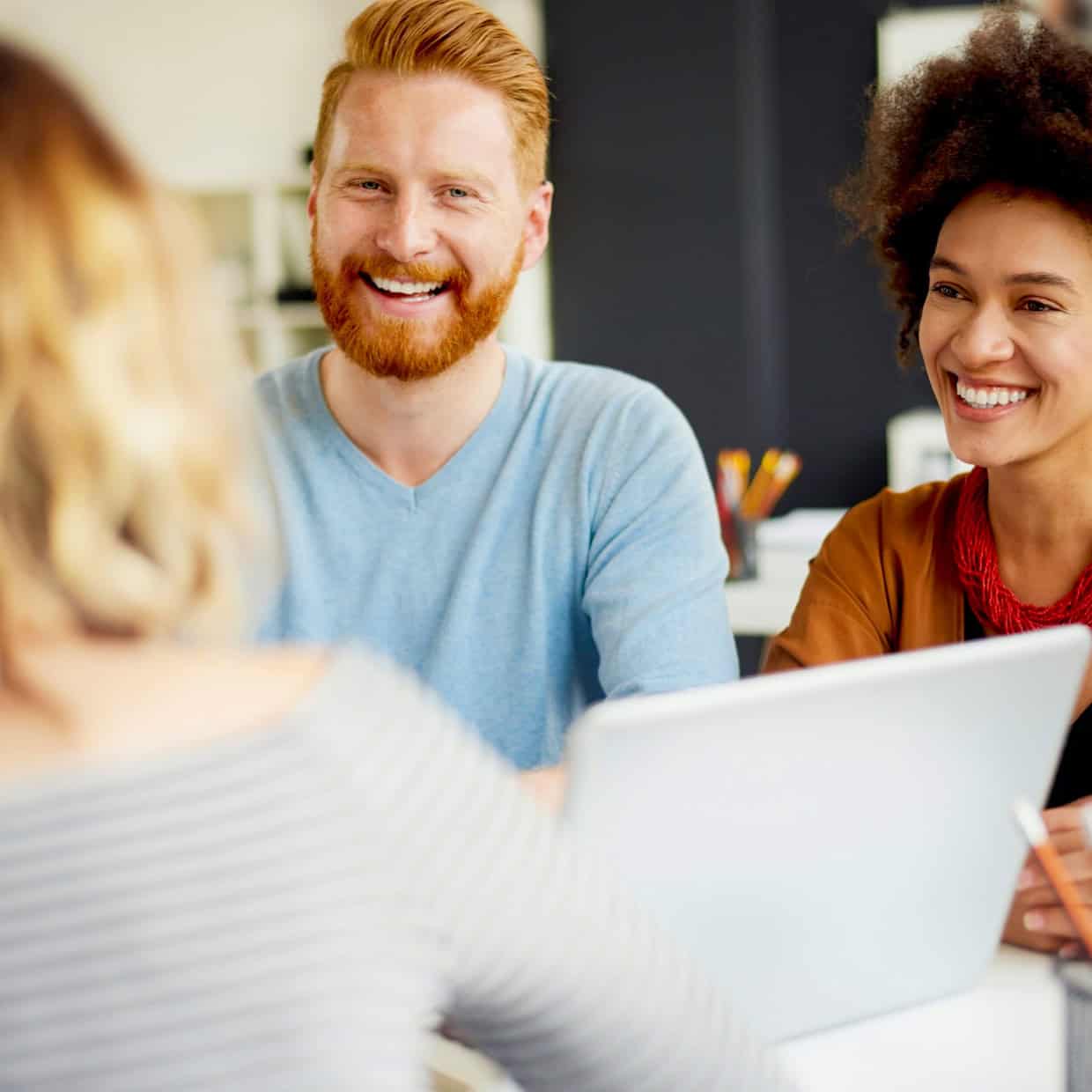 Smiling couple sitting at a desk with a laptop, talking to a person facing them in an office setting.