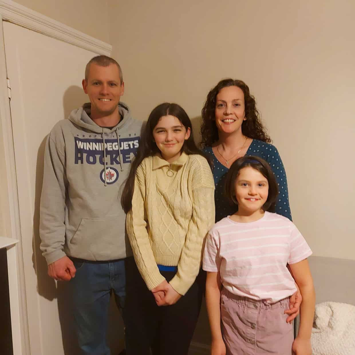 Smiling family of four standing together indoors, with parents behind and two daughters in front near a light-coloured wall and sofa.