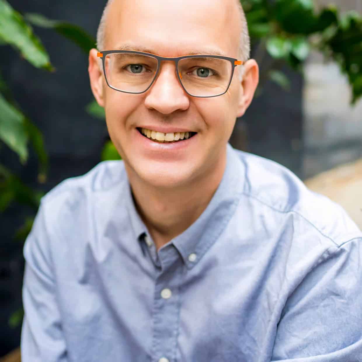 Smiling man wearing glasses and a light blue shirt, seated indoors with green plants in the background.