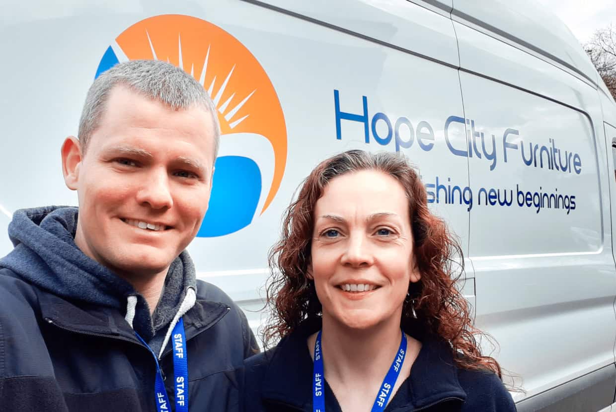 Two staff members smiling in front of a van with the Hope City Furniture logo and the slogan 'Furnishing new beginnings' displayed on the side.