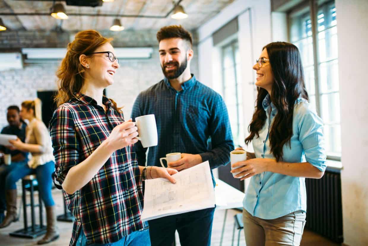 Three co-workers chatting and holding mugs in a bright modern office.