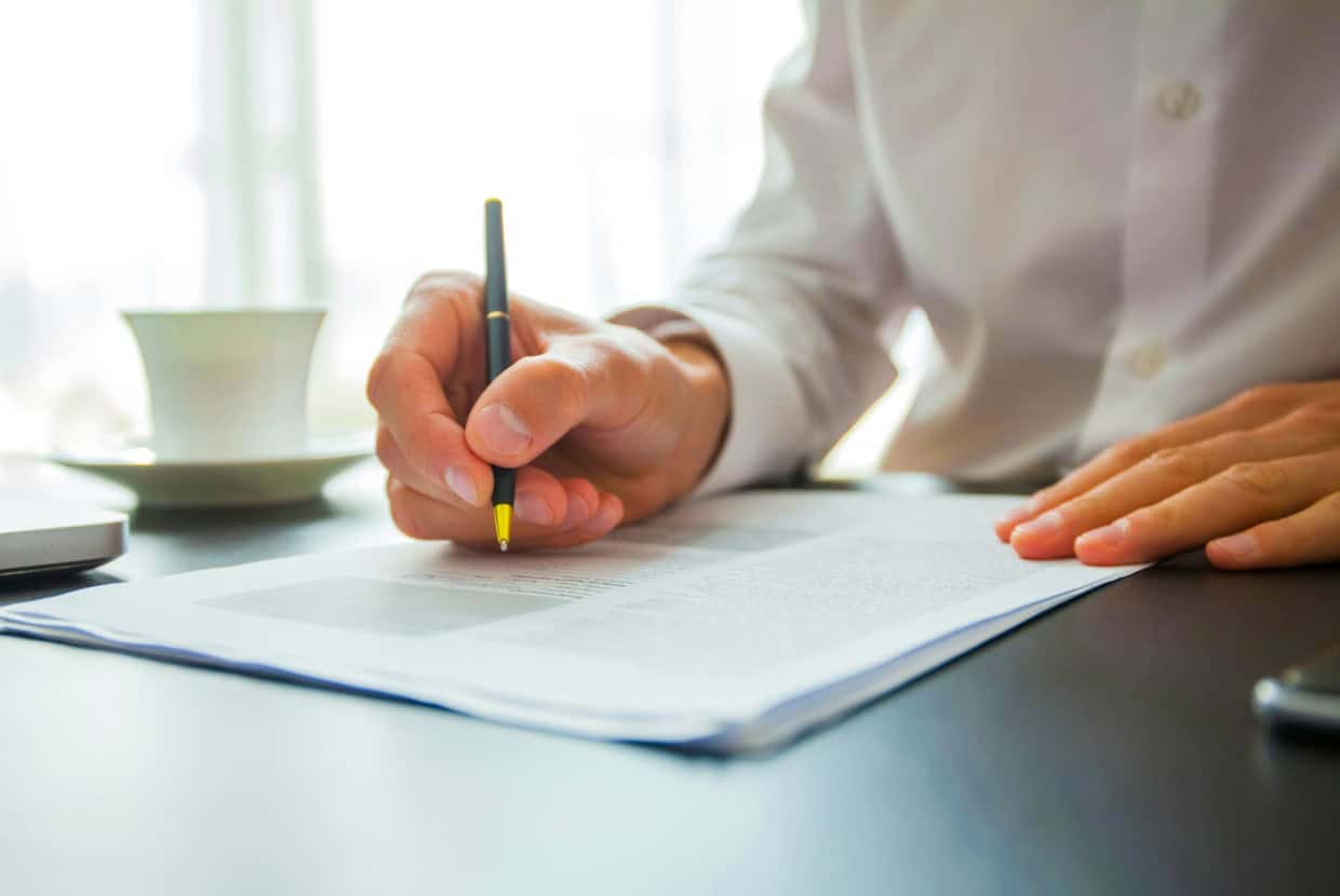 Person in a white shirt signing a document at a desk with a cup and saucer nearby.