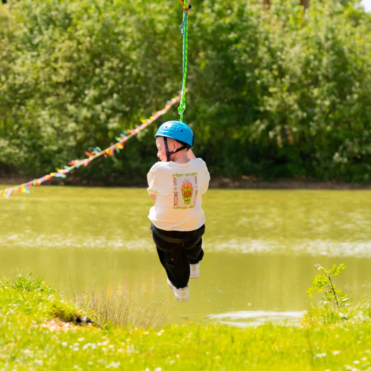 Child wearing a blue helmet and white T-shirt riding a zip line over a small lake surrounded by green trees and grass.