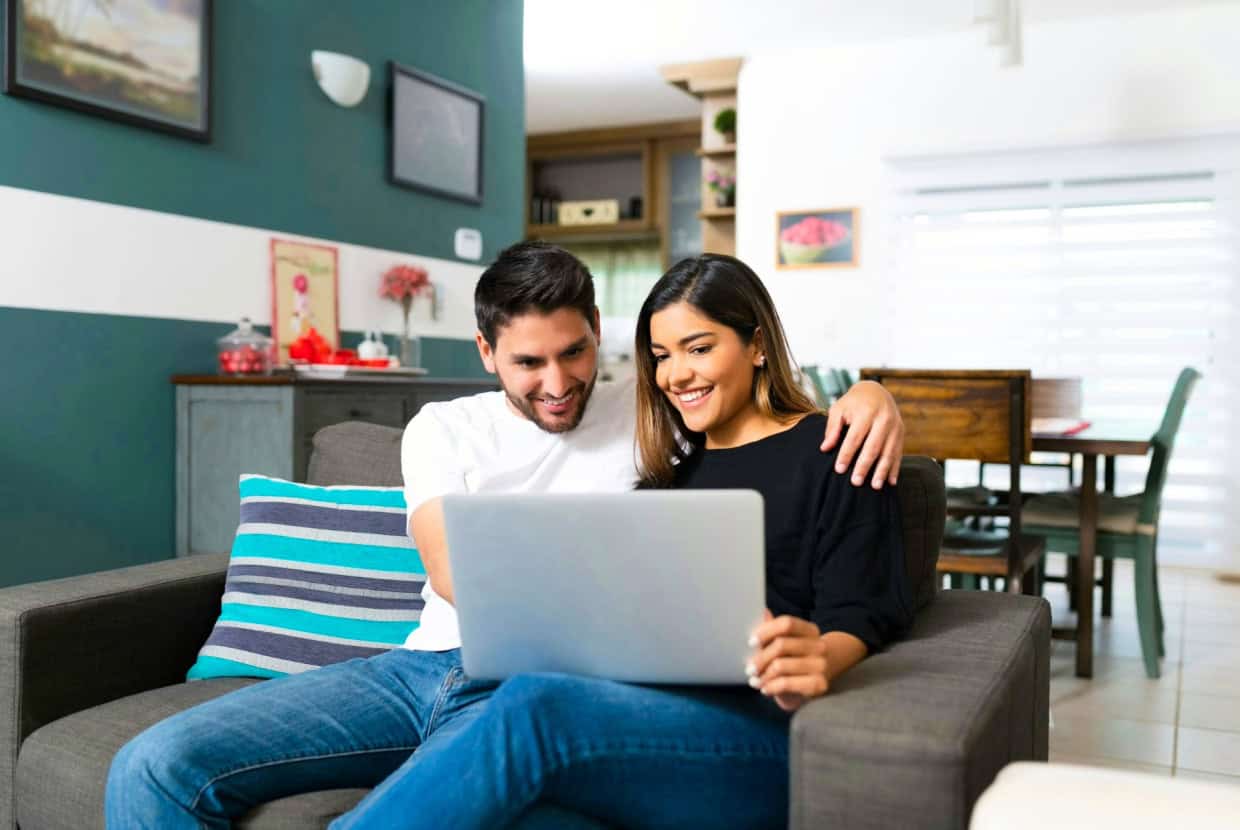 Smiling couple sitting on a sofa at home, looking at a laptop together.