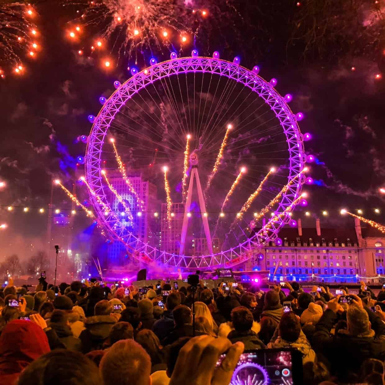 Large crowd watching fireworks bursting around the illuminated London Eye at night during New Year celebration.