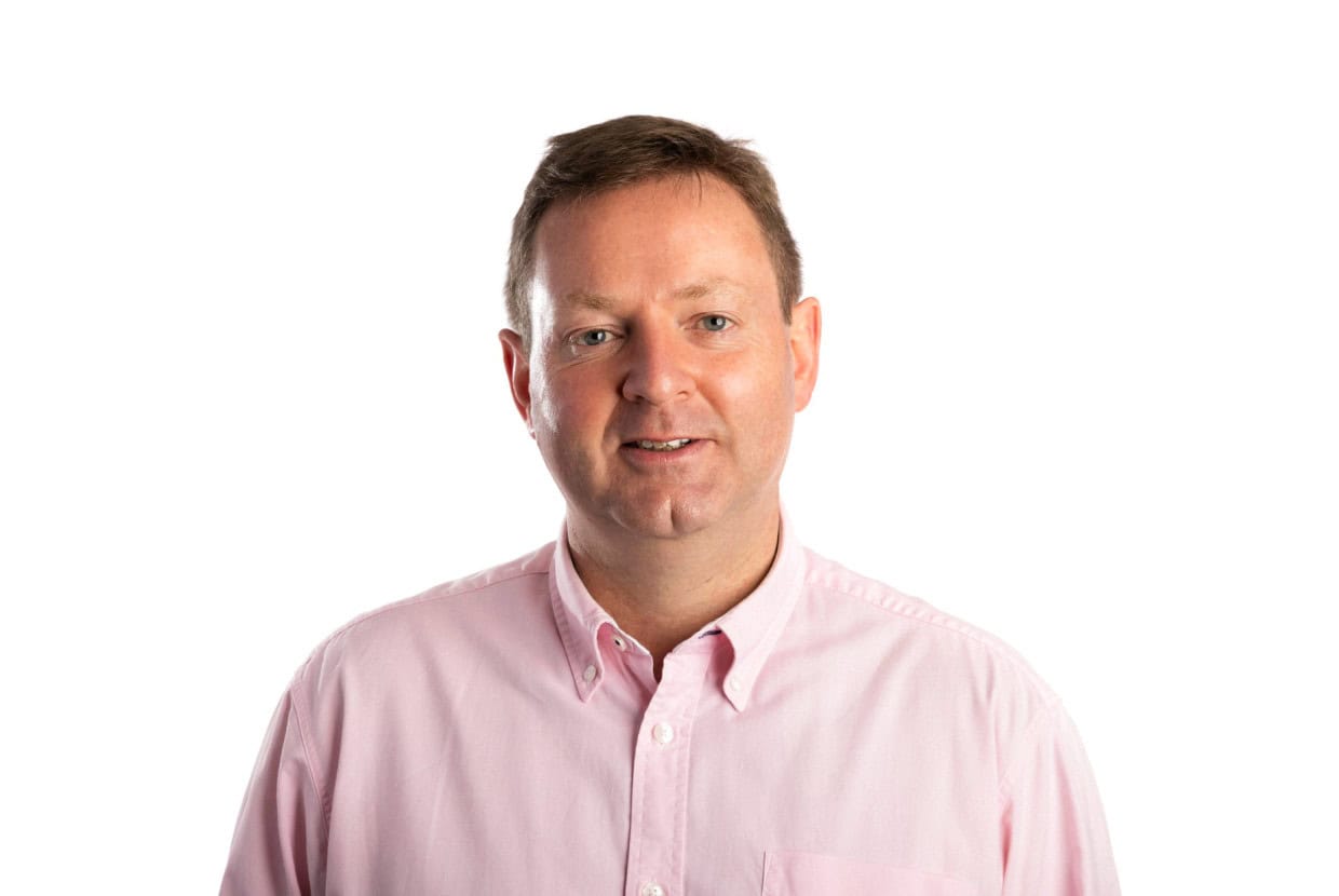 Man wearing a light pink button-up shirt smiling against a plain white background.