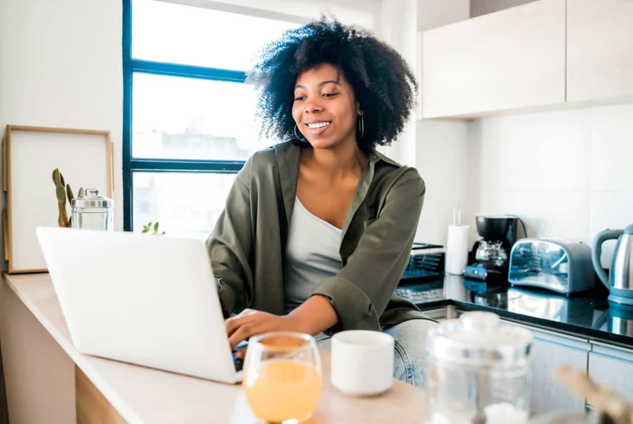 Young woman smiling while working on a laptop in a bright kitchen.