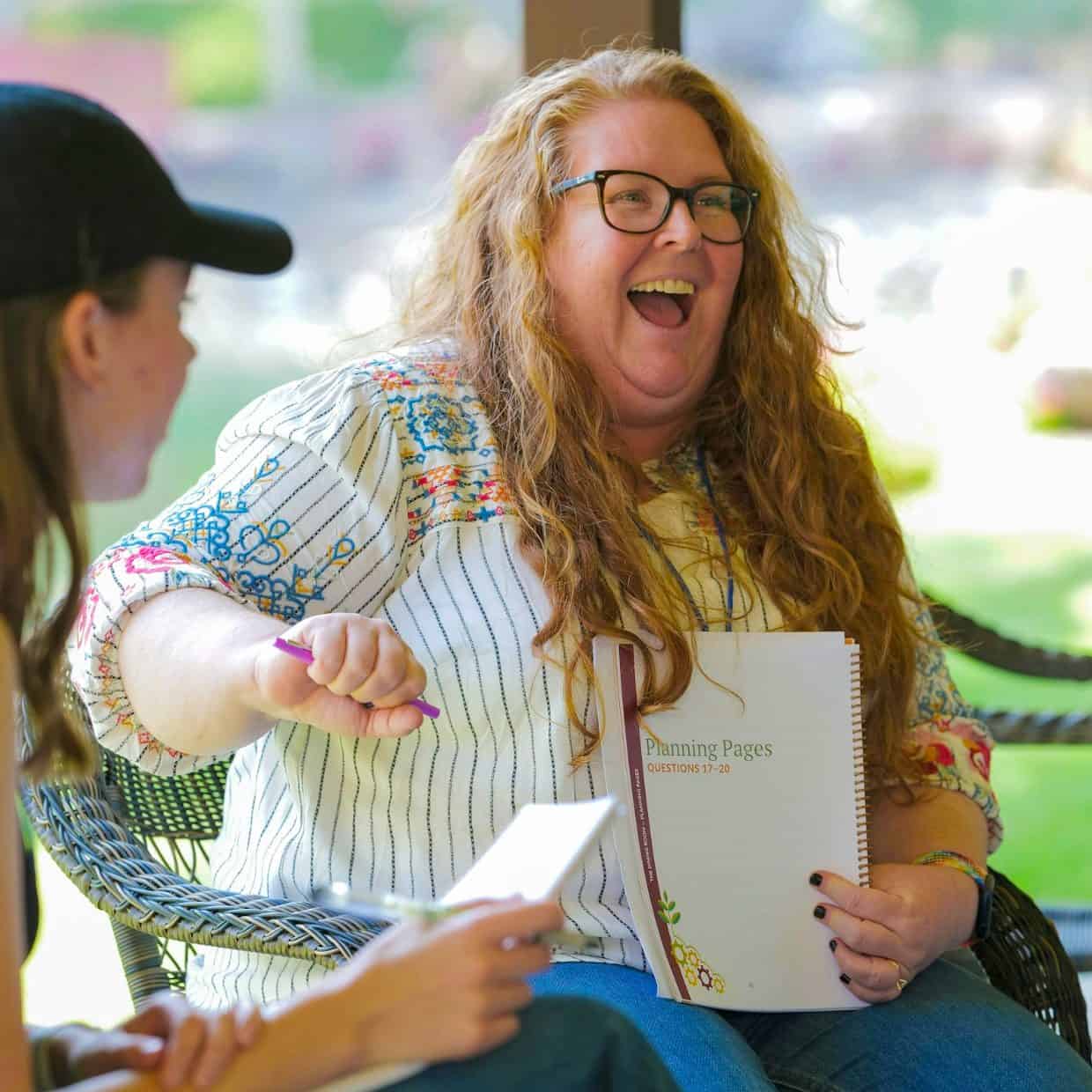Two women laughing and talking outdoors while holding notebooks and pens.