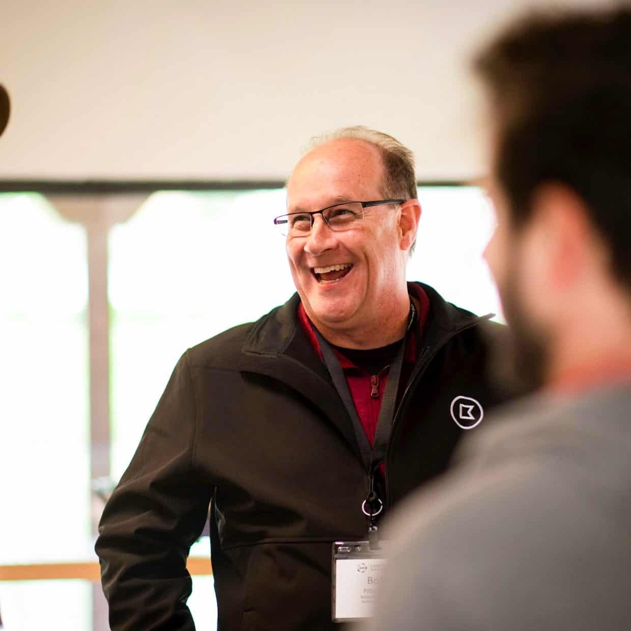 Man in glasses smiling while talking with a group at an indoor event.