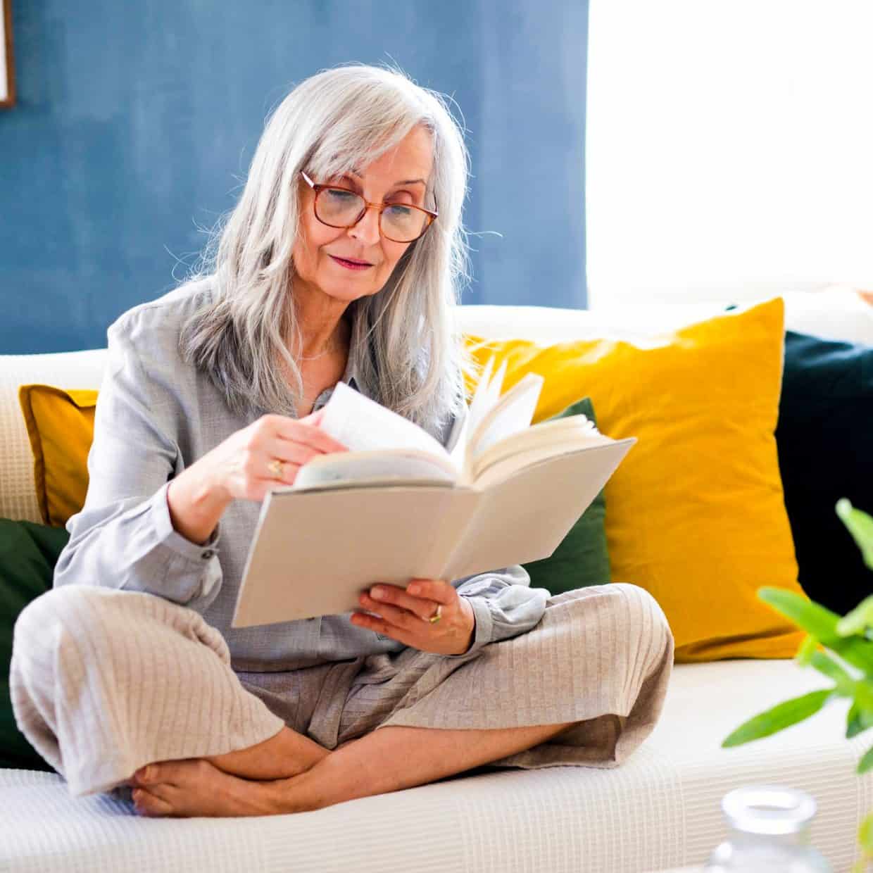 Older woman with grey hair reading a book on a sofa with colourful cushions.