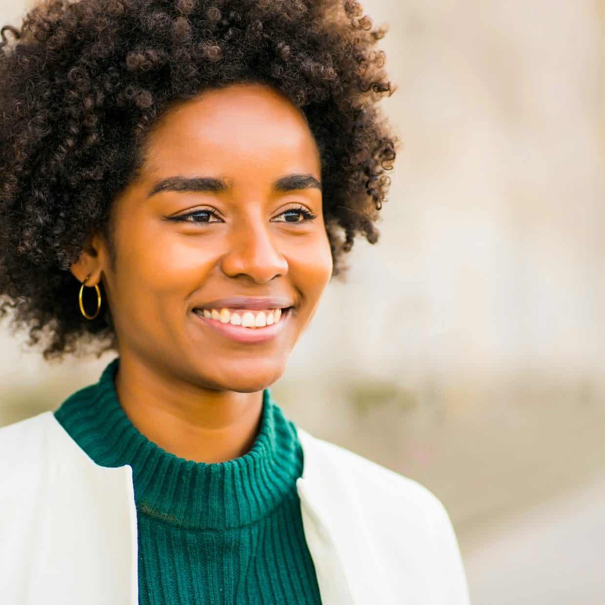 A smiling woman with curly hair wearing a green jumper and white blazer, standing outdoors.