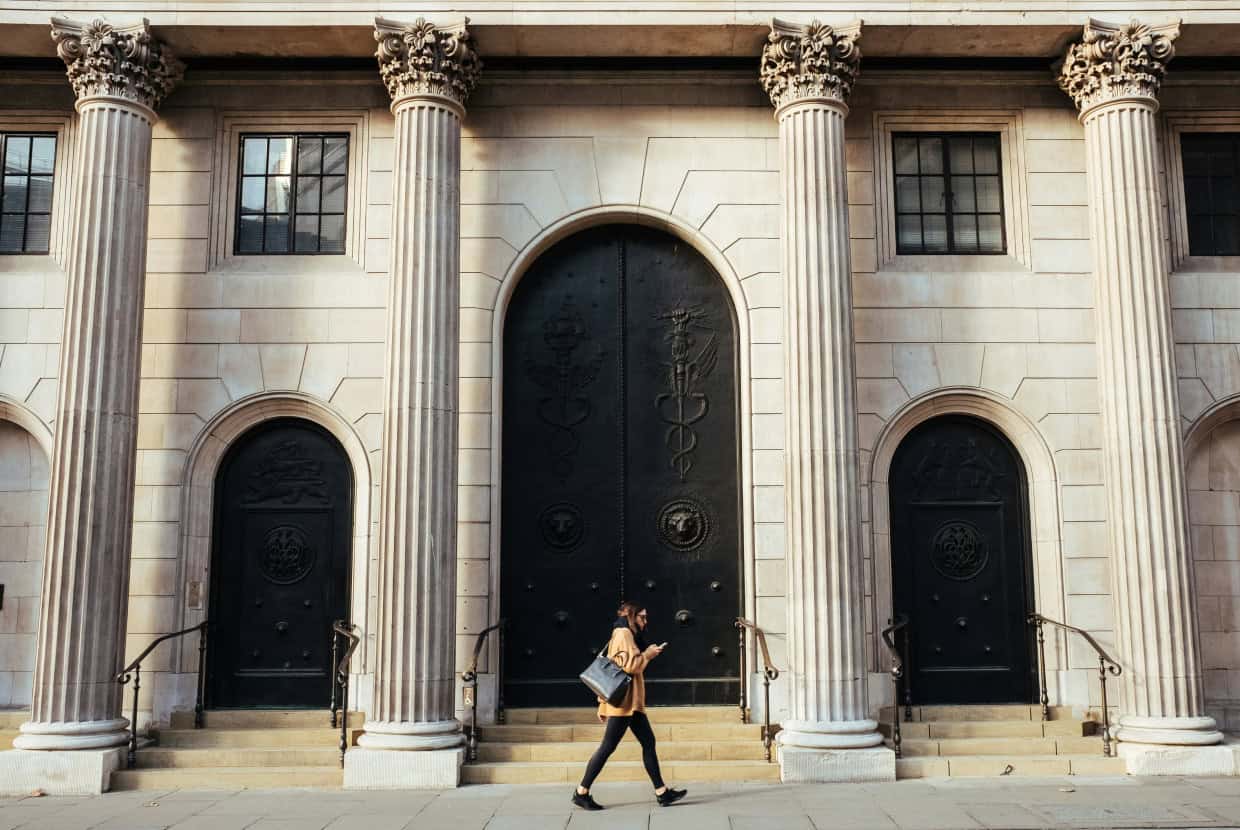 Person walking past a grand neoclassical building with tall columns and large ornate doors, suggesting a historic or financial institution.