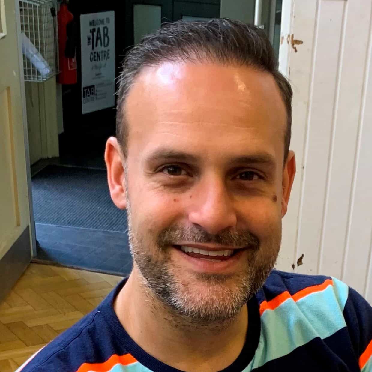 Smiling man with short dark hair and trimmed beard wearing a striped shirt, seated indoors with a sign reading 'The Tab Centre' visible in the background.